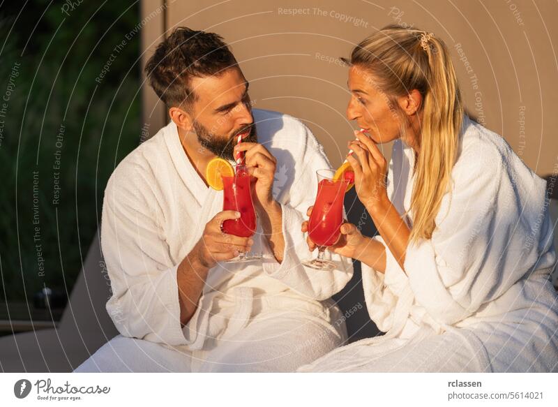 Man and woman in white robes sipping cocktails in golden hour light at spa hotel resort bathrobe wellness spa wellness resort couple red cocktails joyful