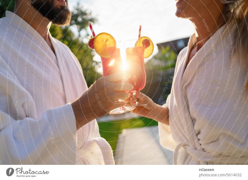 Couple in white bathrobes clinking red cocktails with a sunset behind at spa hotel toasting white robes sunlight outdoors cheers relaxation leisure poolside