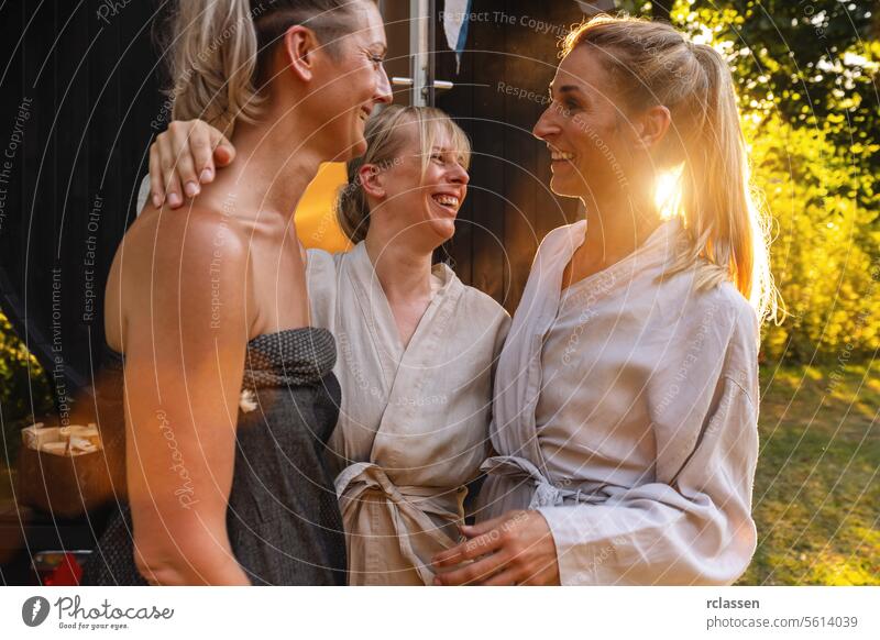 Three happy people in robes by a Finnish sauna barrel, laughing and chatting mobile finnish sauna relaxation wellness friendship bonding spa nature outdoor