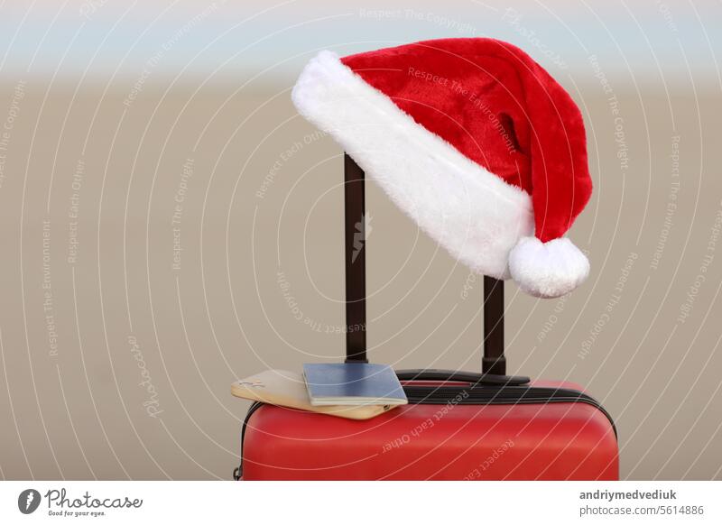 Santa Claus hat, smartphone and passport on red suitcase on seashore sand beach. Travel on Christmas and New Year winter holidays concept. Last minute trips, hot tours. Copy space for advertising