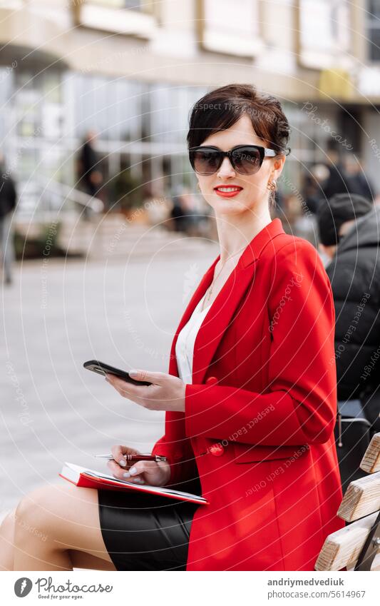 Lawyer successful businesswoman in red blazer and sunglasses is sitting on bench on city street talking on smartphone. Smiling woman making business call on cell and doing notes in notebook outdoors.
