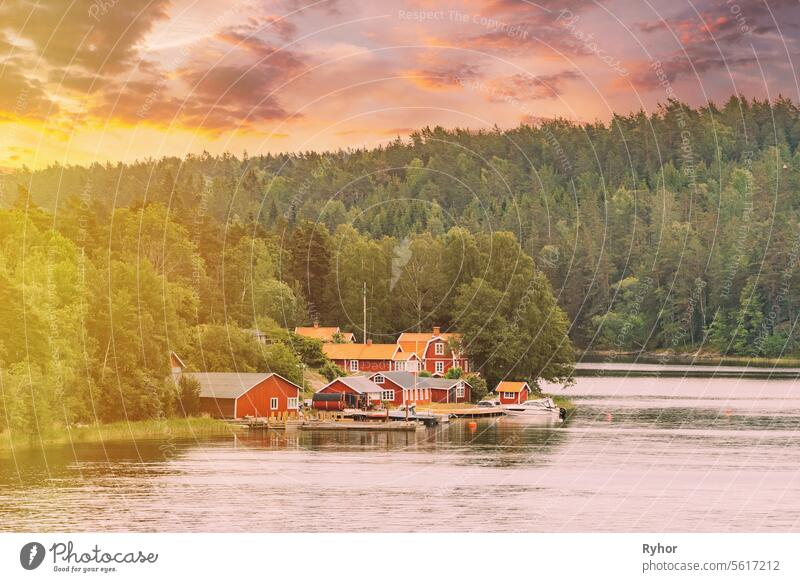 Sweden. Beautiful Red And Yellow Swedish Wooden Log Cabins Houses On Rocky Island Coast In Summer Evening. Lake Or River Landscape. sunset summer sky with sunlight sunshine. altered sunrise sky.