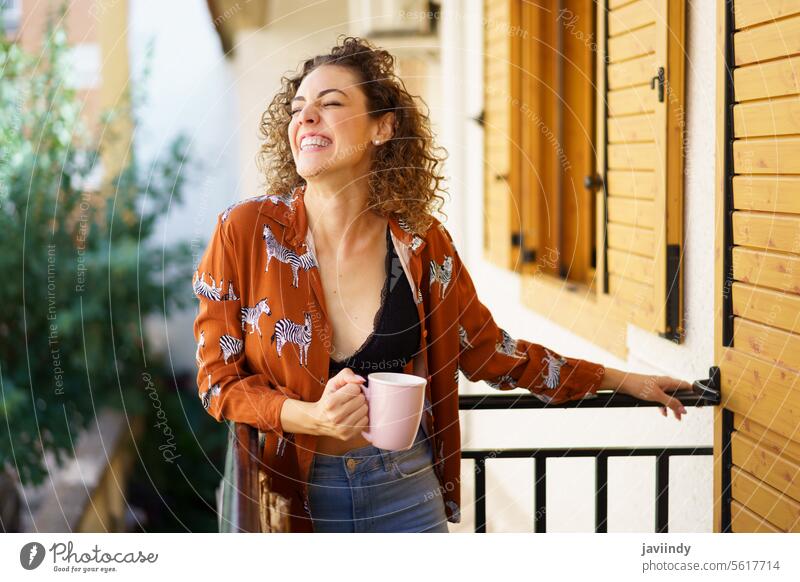 Cheerful young woman holding coffee cup in hand while leaning on balcony of happy style smile trendy cheerful terrace brunette enjoy curly hair outfit positive