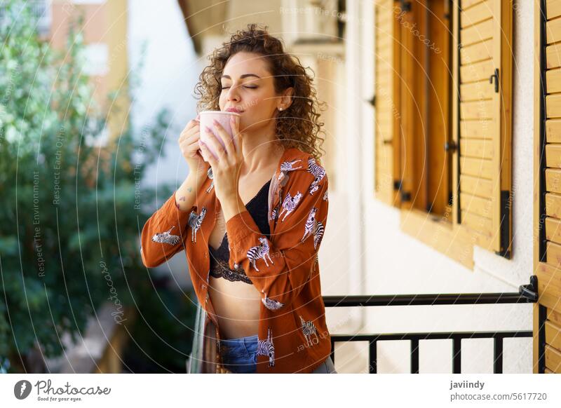 Young woman enjoying coffee while standing on balcony of house tea curly hair building fresh modern pleasure lady drink hot drink brunette street alone pleasant