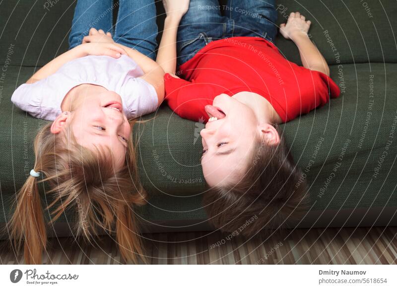 Brother and sister wearing casual clothes laying on a green sofa at home stick out tongues teasing each other Half-sibling boy brother caucasian child childhood