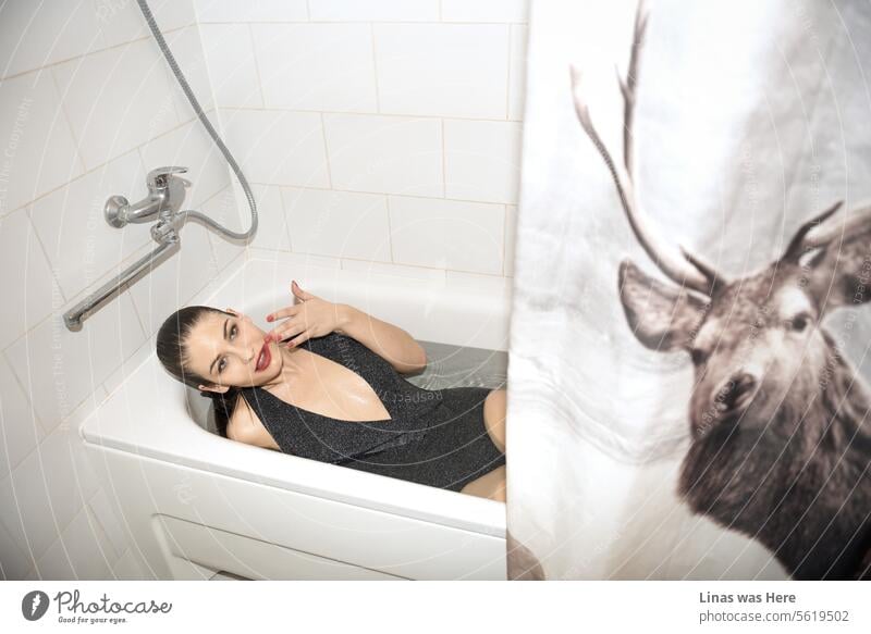 A stunning brunette is enjoying a bath in a tub, adorned in a shiny swimsuit. Her makeup is beautifully done, accentuating her features, and she sports striking red lips. A pretty woman and deer curtains in a bathroom.
