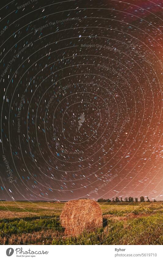 Night Starry Sky in rotation Above Country Field Meadow With Hay Bale After Harvest. Glowing Stars Above Rural Landscape. Colorful flare on sky. Agricultural Landscape under Starry Sky. Colored bright spots of light glow in the sky. sky rotate. Stars Ba...