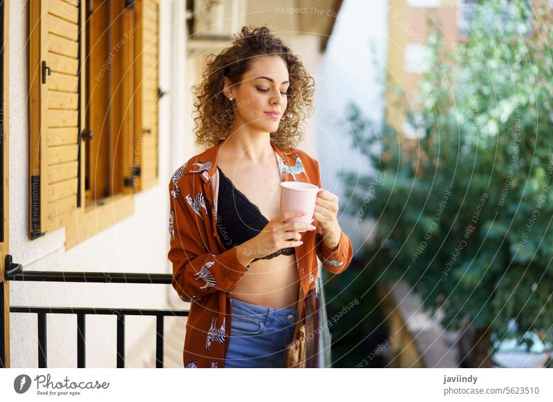 Young stylish smiling woman with cup of coffee standing on balcony at home smile fashion drink outfit curly hair enjoy confident content glad optimist gorgeous