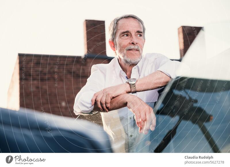 Smiling elderly gentleman in white shirt casually leaning on his convertible looking into the distance, in front of an old industrial backdrop Best Ager