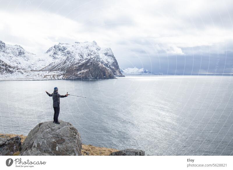 man in winter landscape, sheltered man with open arms standing on a rock in front of the dark sea, in the background you can see the snow-capped mountains, it is cold and the sky is cloudy