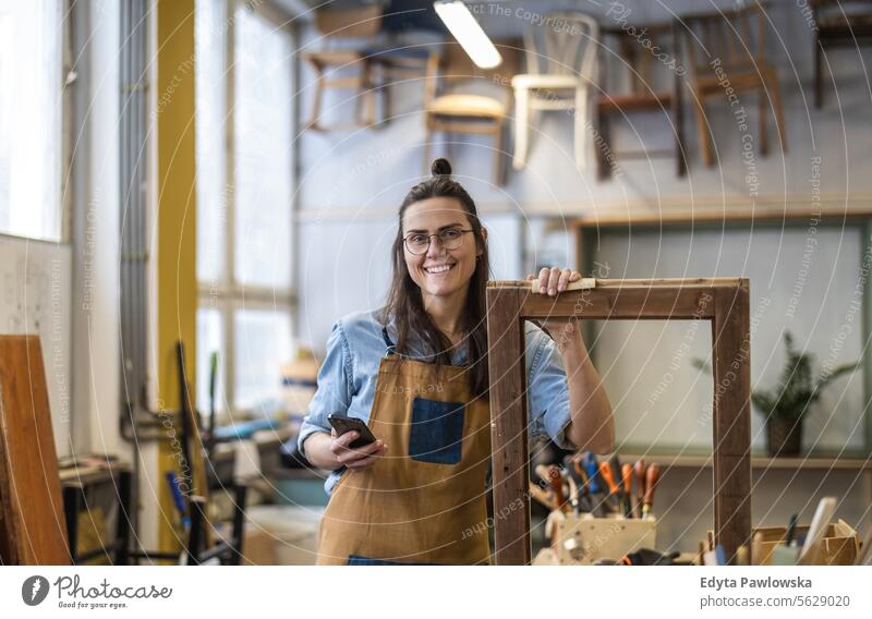 Portrait of young female carpenter using mobile phone in her workshop Furniture Carpenter Restoring Carpentry wood Chair building Manufacturing Craft Renovation