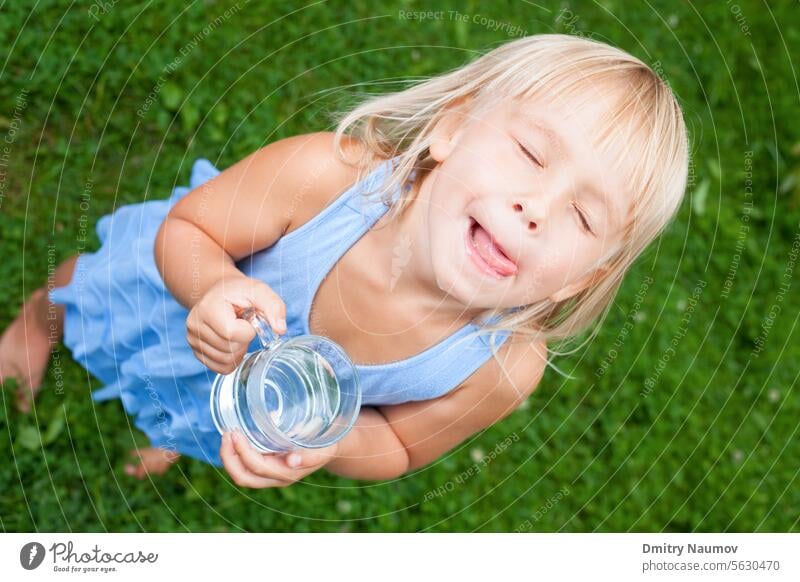 High angle view shot of blonde little girl wearing blue dress holding glass of water licking her lips with her eyes closed in a summer garden 4-5 years aqua