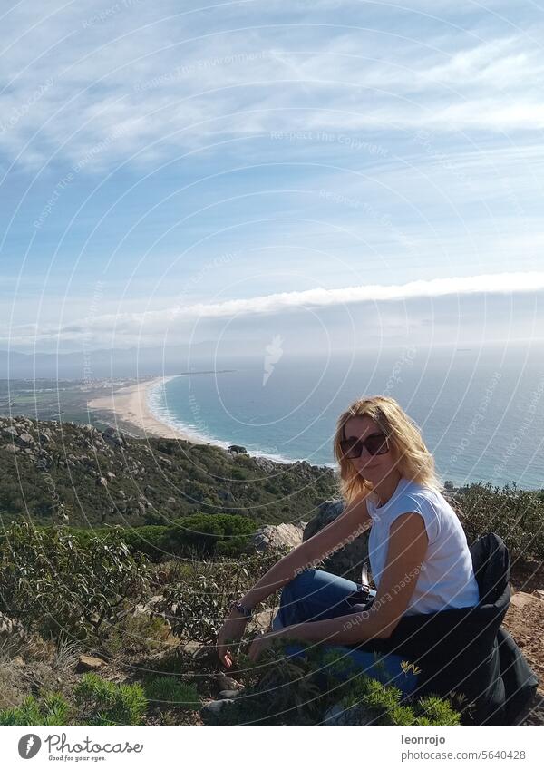 A beautiful woman in a T-shirt and sunglasses sits on a mountain during the day and enjoys the peace and quiet and the panorama. In the background you can see a long beach and the ocean.