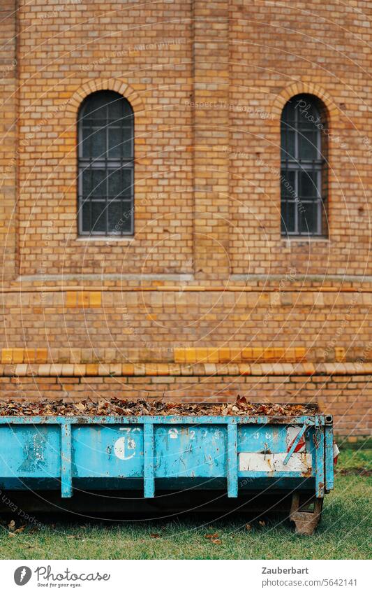 Light blue container with building rubble stands in front of a church with a brick wall and two arched windows Container light blue Building rubble Church duo