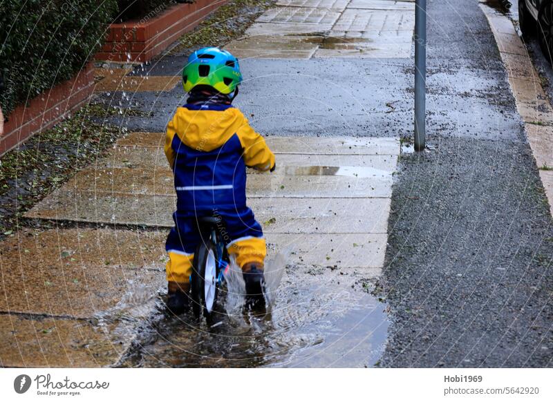 Small child riding a balance bike through a puddle Child Puddle impeller Wheel Walking Bicycle Cycling Water Inject Rain fun Rain jacket Jacket Helmet
