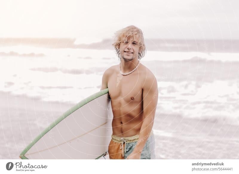 Handsome fit young blond man with mock up surfboard waits for wave to surf spot at sea ocean beach with black sand and looks at camera. Concept of sport, fitness, freedom, happiness, new modern life