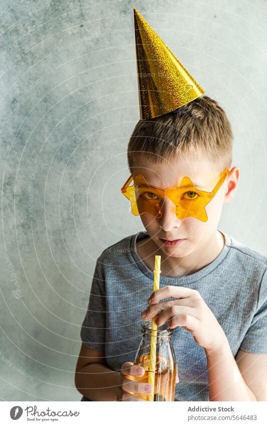 Boy in party hat and star-shaped glasses sipping a drink boy new year's glass bottle straw birthday celebration glittery yellow neutral backdrop child festive