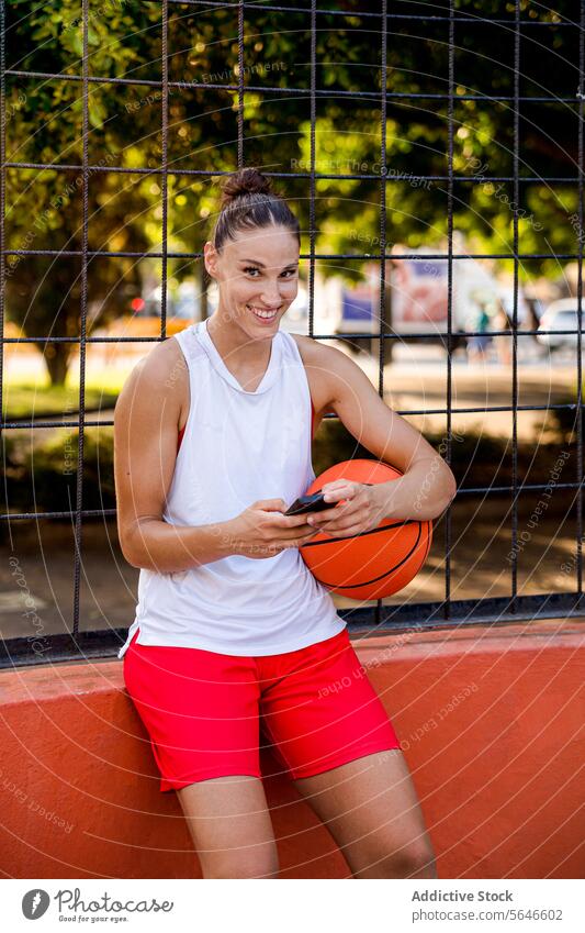 Smiling young female basketball player holding ball and using smartphone while looking at camera in playground on sunny day Sportswoman Using Smartphone