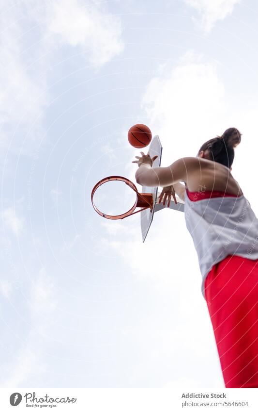 From below of cropped unrecognizable female basketball player in activewear jumping and throwing ball into basket hoop against cloudy sky Sportswoman Basketball