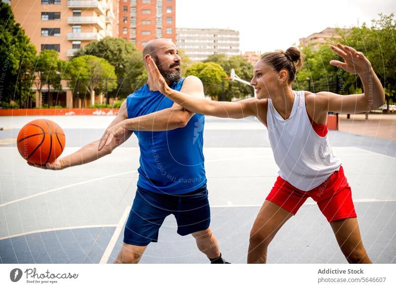 Determined man and woman in sportswear playing basketball on playground in city with buildings and trees in background Sportspeople Play Basketball Ball Defend