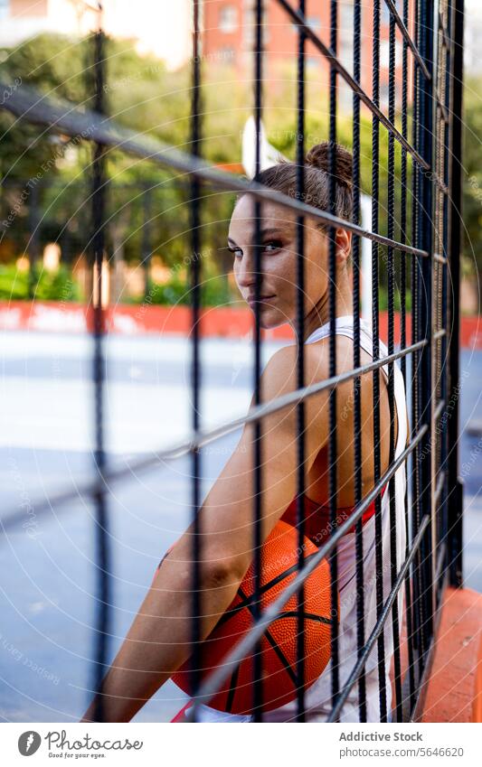 Confident young female athlete in sportswear holding basketball looking at camera seen through fence of playground Sportswoman Basketball Ball Athlete Player