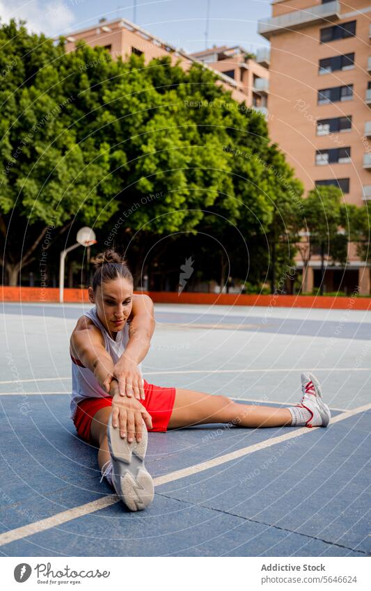 Full body of determined young female basketball player sitting and touching feet while doing warmup exercise on playground Sportswoman Basketball Player Warm Up