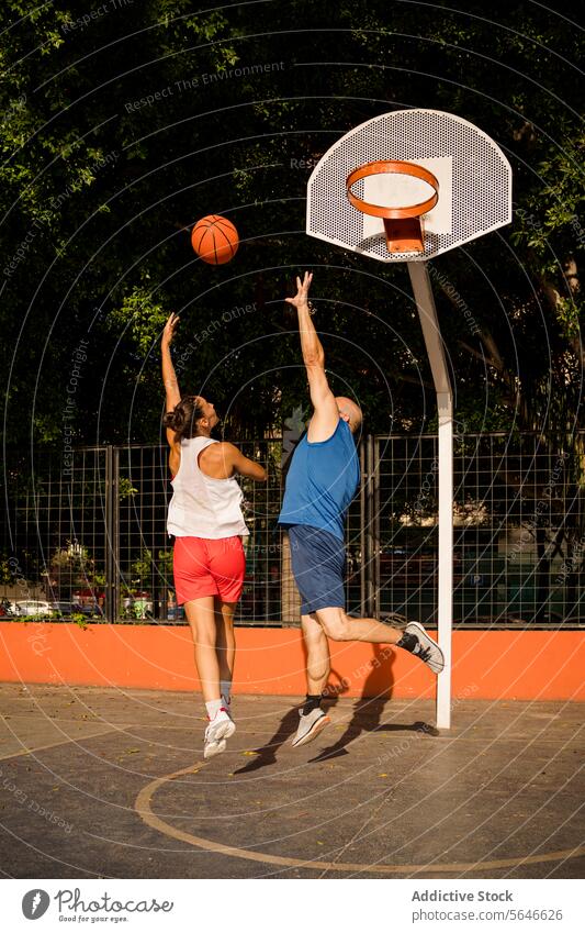 Full length back view of sportswoman in activewear shooting basketball towards basket and man trying to defend on court against sky Sportspeople Basketball
