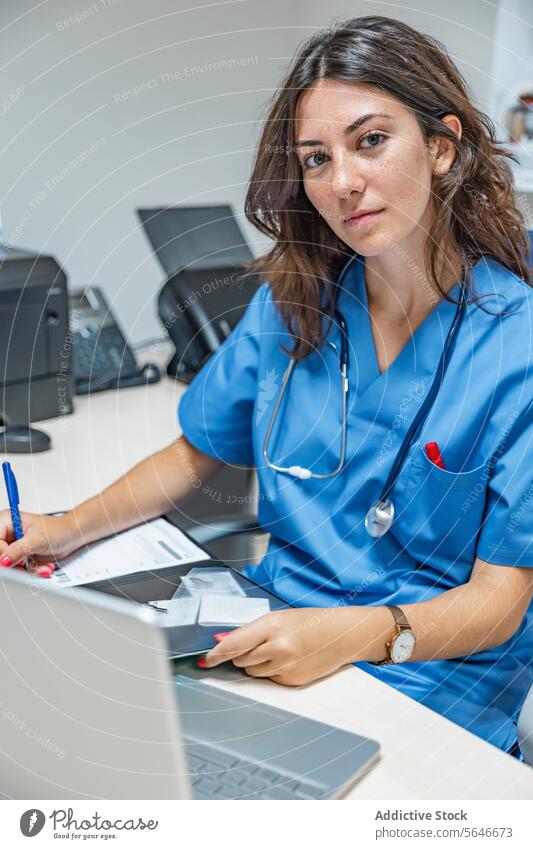 Young woman doctor sitting at table with laptop smartphone and making note on clipboard write smile stethoscope pen clinic female young professional specialist