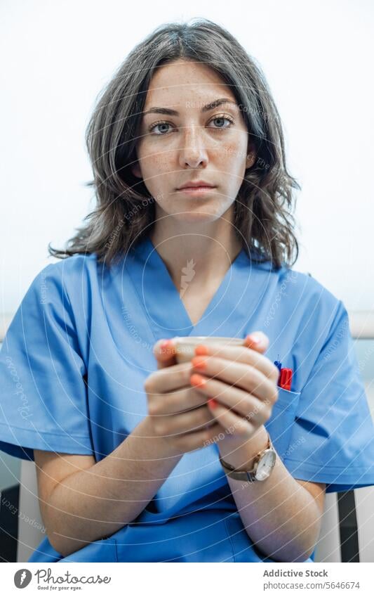 Young female doctor holding cup of coffee and looking at camera while sitting woman uniform hospital medical health care specialist young professional medicine
