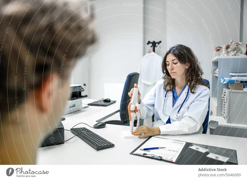 Woman doctor sitting at table with tendon and muscle figurine in clinic and showing to unrecognizable man patient woman uniform stethoscope clipboard hospital