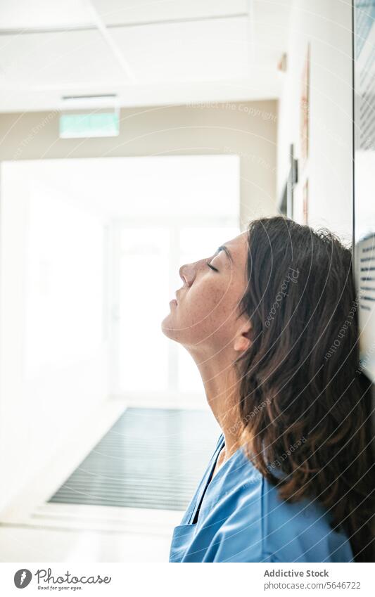 Young female doctor relaxing while leaning against wall in hospital woman eyes closed clinic medicine uniform curly hair treat modern health care specialist