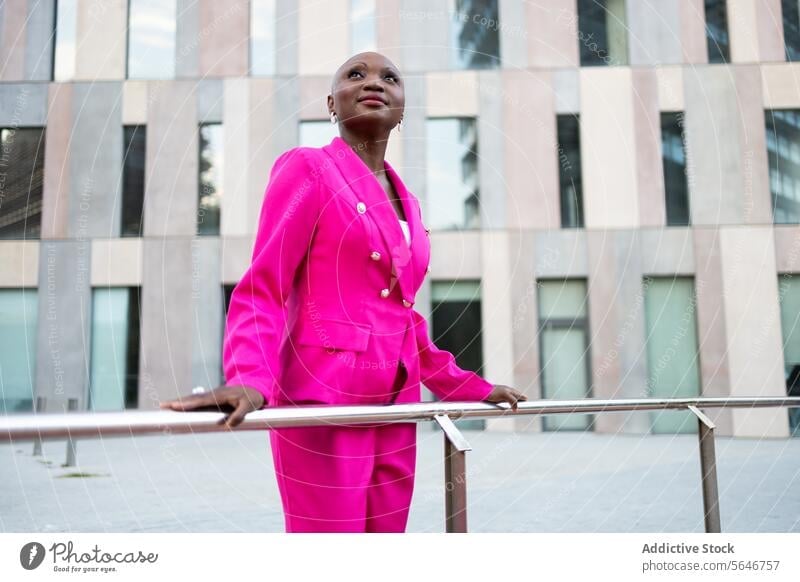 Smiling Elegant black woman with shaved head in pink outfit standing on street against building trendy look style appearance dreamy jacket elegant personality