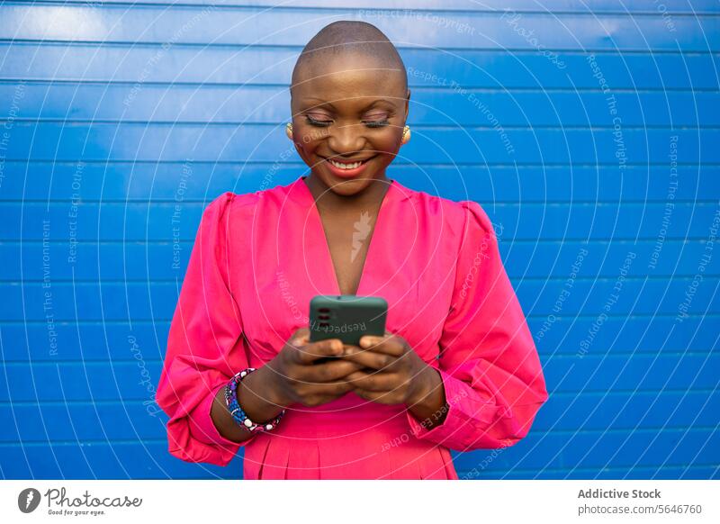 Cheerful black woman with shaved head in pink clothes using smartphone against blue wall positive browsing dress smile happy text message cheerful outfit mobile