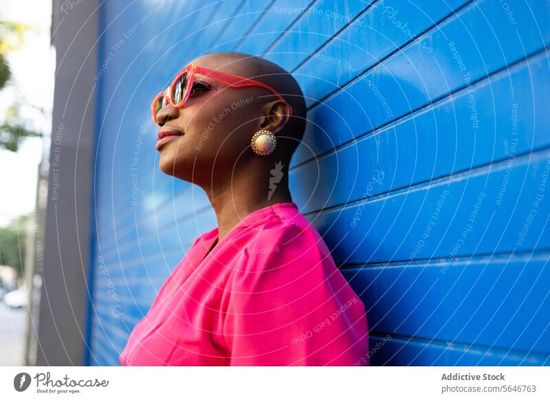 Happy Stylish black woman with shaved head in sunglasses and pink dress standing against blue wall confident cool trendy outfit individuality style smile happy