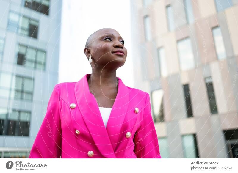 Low angle of Elegant black woman with shaved head in pink outfit standing on street against buildings trendy look style appearance dreamy jacket elegant