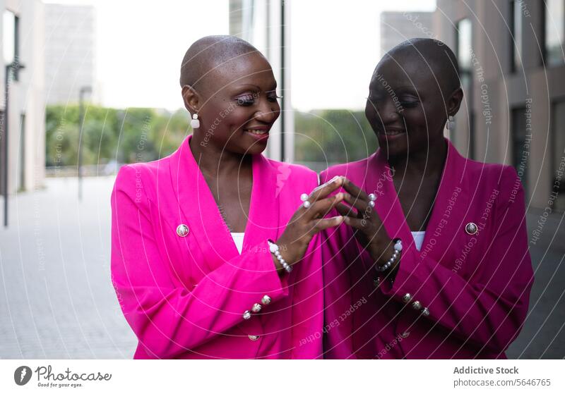 Cheerful black woman with shaved head in pink outfit with reflective mirror image in daylight style street suit makeup reflection appearance glass wall elegant