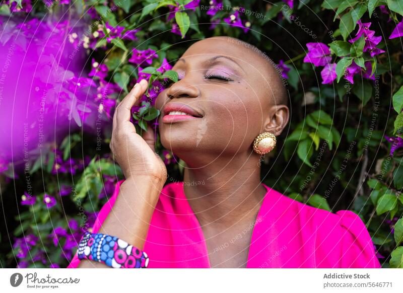 Happy black woman with shaved head in pink dress with closed eyes standing near blooming bush in park bougainvillea flower purple plant smile nature positive