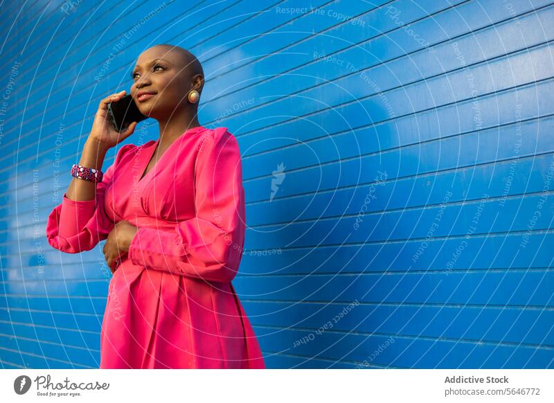 Stylish black woman with shaved head in pink dress talking on cellphone against blue wall smartphone using phone call happy bald conversation speak smile ethnic