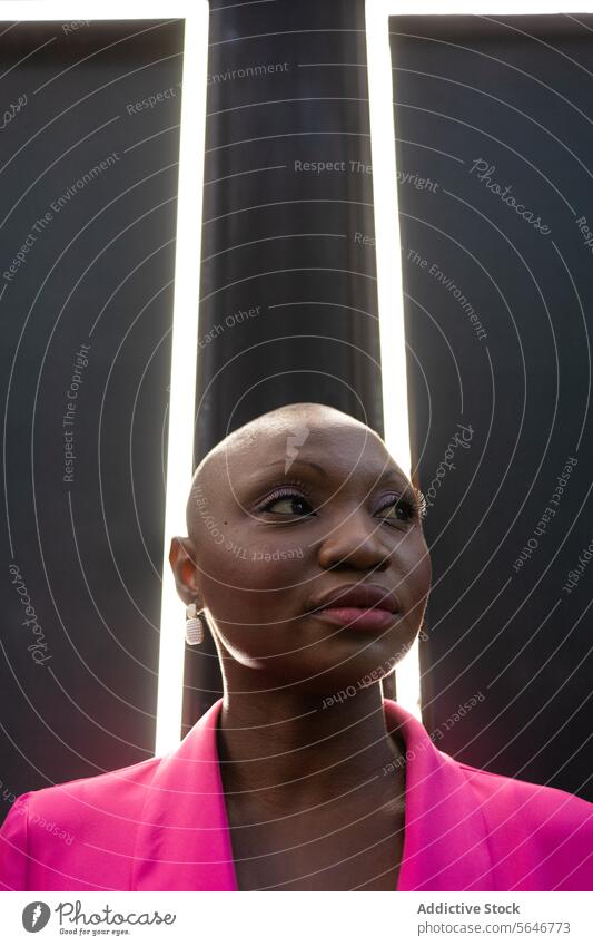 Confident black woman with shaved head in pink outfit standing in front of frames with lights confident wall individuality appearance earring posture summer
