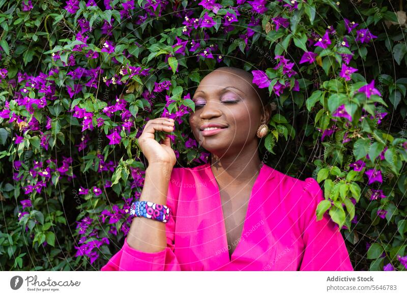Happy black woman with shaved head in pink dress with closed eyes standing near blooming bush in park bougainvillea flower purple plant smile nature positive