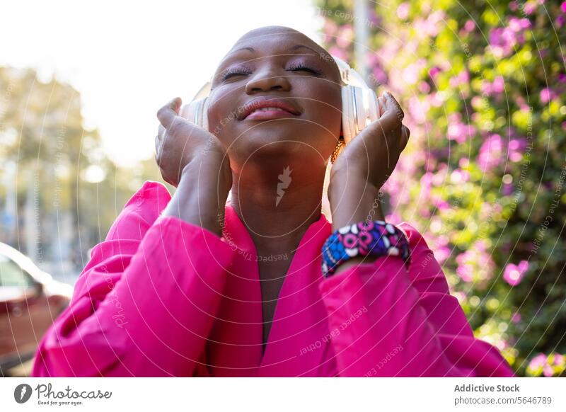 Happy black woman with shaved head in pink dress listening to music through headphones with closed eyes in park wireless using happy carefree trendy style