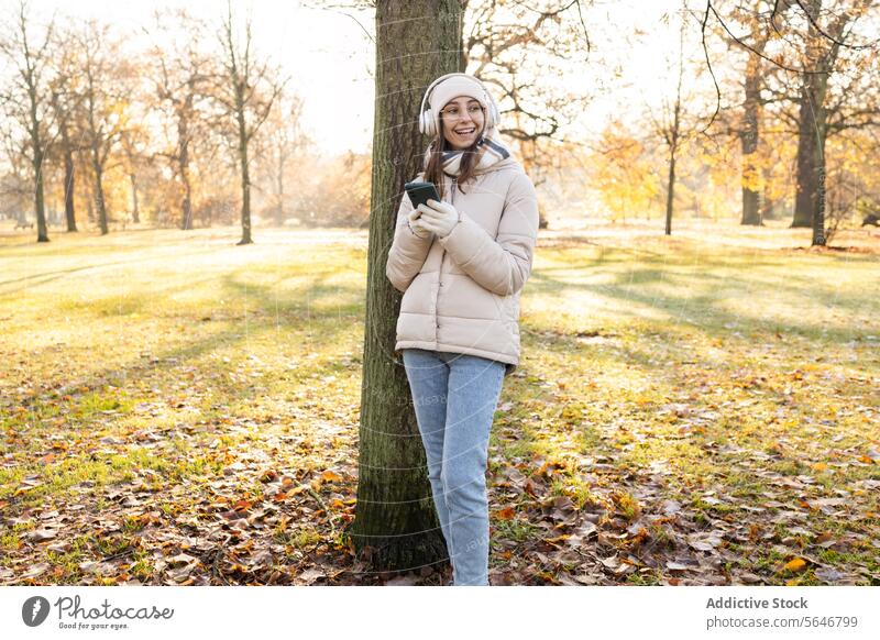 Cheerful young woman in warm clothes using smartphone in park listen music headphones autumn positive smile female mobile happy device enjoy hat cellphone