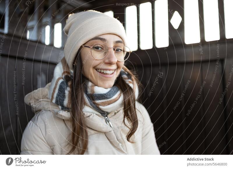 Smiling young woman in warm jacket standing by wooden fence smile winter autumn warm clothes positive happy hat portrait eyeglasses glad cap delight surfing