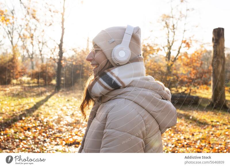 Smiling woman in headphones listening to music in autumn park smile song positive enjoy leisure warm clothes glasses wireless female young happy eyeglasses hat