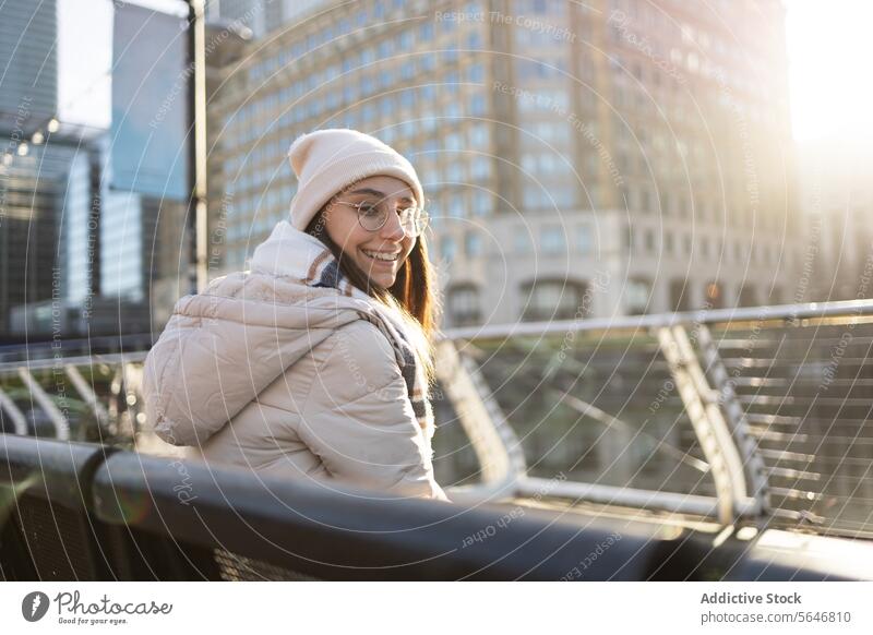Smiling woman in warm clothes sitting on metallic bench by railing in city street building modern positive young female cheerful urban smile glad lifestyle