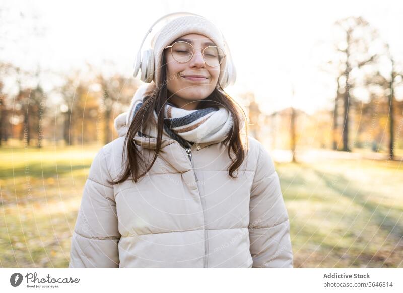 Smiling woman in headphones listening to music in autumn park smile song positive enjoy leisure warm clothes glasses wireless female young happy eyeglasses hat
