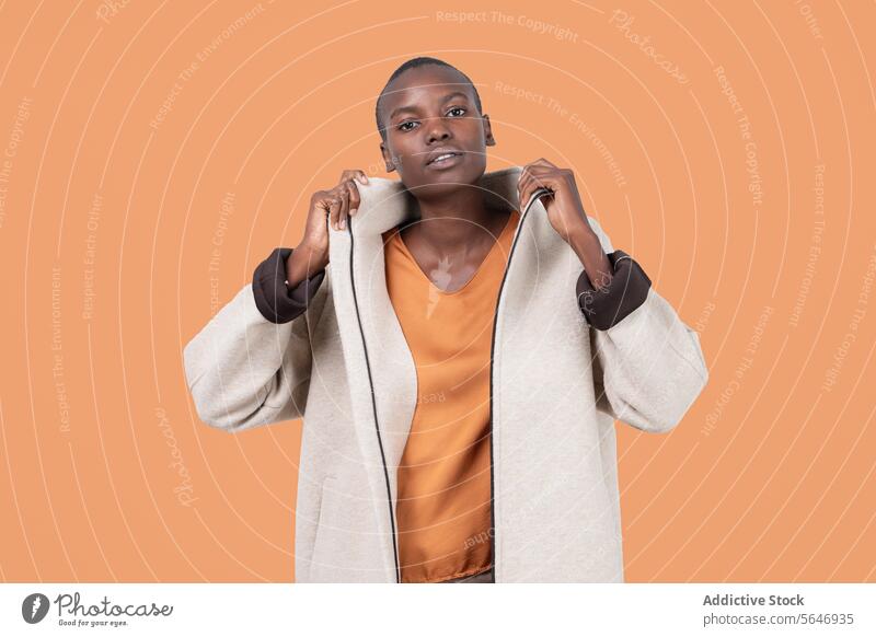 Portrait of young bold African American female in long zipper coat confidently looking at camera standing on orange background Woman Confident Coat Long Zipper