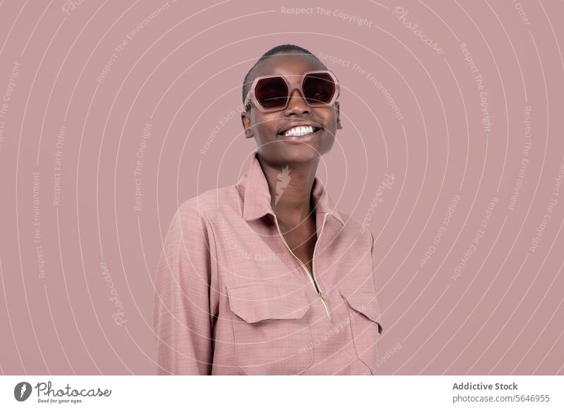 Young cheerful bold African American female model in stylish pink clothes and sunglasses standing looking away against pink background Woman Style Trendy Outfit