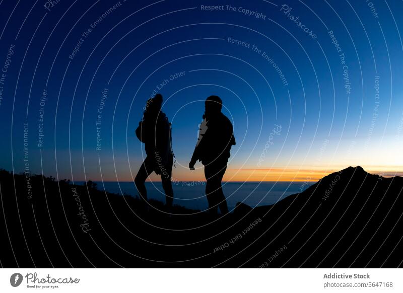 Silhouette of unrecognizable traveler couple admiring orange sunset on highland cliff silhouette mountain admire blue sky nature male female stand trip journey