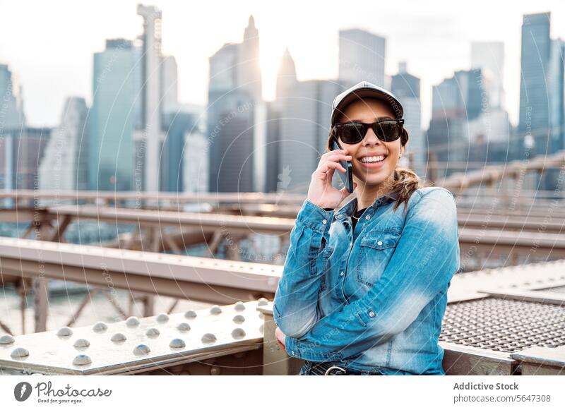 Woman chatting on phone against Manhattan view woman call sunglasses skyline cityscape New York conversation urban bridge fashion joy denim glow lifestyle